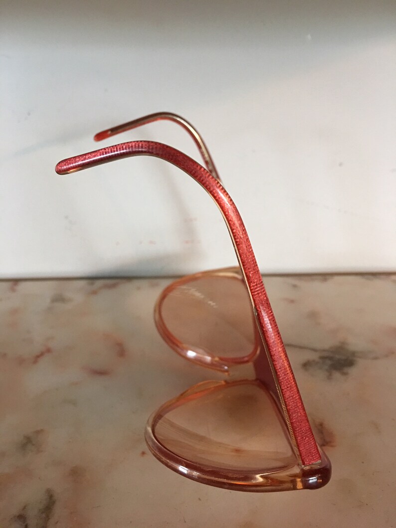 May include: A pair of pink plastic sunglasses with red frames. The sunglasses are in a side view, with the arms of the glasses folded up.