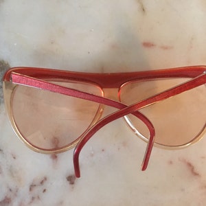 May include: Red and peach plastic sunglasses with a cat-eye shape. The sunglasses are on a white marble surface.