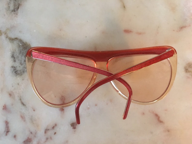 May include: Red and peach plastic sunglasses with a cat-eye shape. The sunglasses are on a white marble surface.