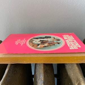 May include: A pink paperback book with a circle on the cover featuring a group of people. The title of the book is "The Pump House Gang" by Tom Wolfe.