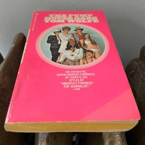 May include: A pink paperback book with a yellow spine. The cover features a group of people in a circle, with the title "The Pump House Gang" and the author "Tom Wolfe" in white text.