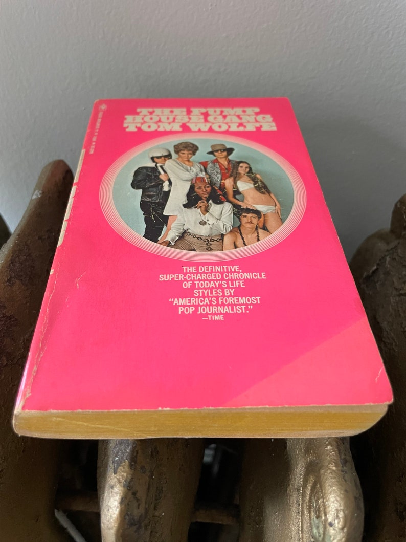 May include: A pink paperback book with a yellow spine. The cover features a group of people in a circle, with the title "The Pump House Gang" and the author "Tom Wolfe" in white text.