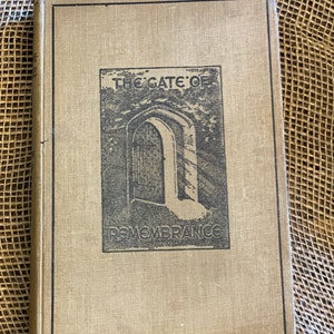 May include: A vintage book titled "The Gate of Remembrance" with a beige cover and black border. The cover features a black and white illustration of an arched doorway. The book is resting on a textured, neutral-toned surface.