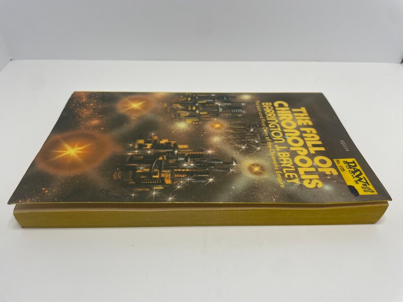 May include: A paperback book cover with a dark background and a cityscape with stars. The title of the book is "The Fall of Chronopolis" by Barrington J. Bayley. The subtitle is "The Last and First Days of the Chrenatic Empire".