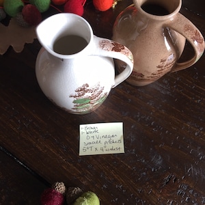 Puede incluir: Dos jarras de cer&aacute;mica vintage, una marr&oacute;n y otra blanca, con un dise&ntilde;o floral. Las jarras miden 14 cm de alto y 10 cm de ancho. Una nota manuscrita dice: 1-Brown 1-White 6 TVinegar Small pitchers 5 1/2" x 4" widest