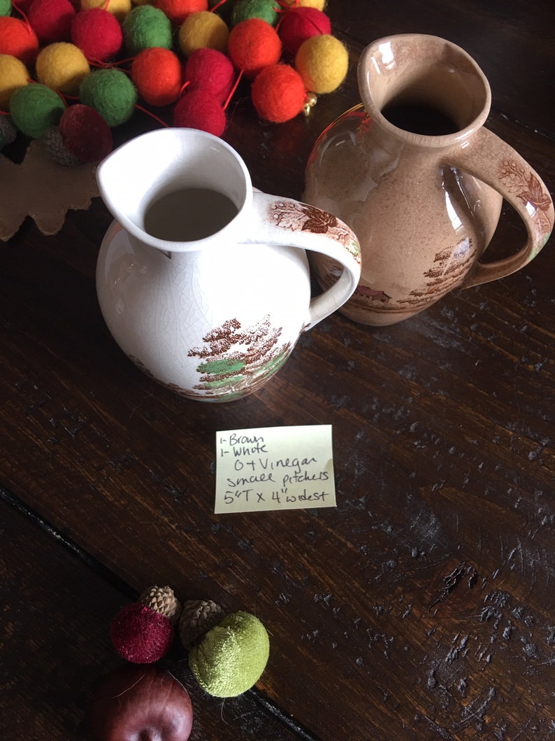 Puede incluir: Dos jarras de cer&aacute;mica vintage, una marr&oacute;n y otra blanca, con un dise&ntilde;o floral. Las jarras miden 14 cm de alto y 10 cm de ancho. Una nota manuscrita dice: 1-Brown 1-White 6 TVinegar Small pitchers 5 1/2" x 4" widest