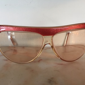 May include: A pair of vintage sunglasses with a red frame and clear lenses. The frame has a textured design.