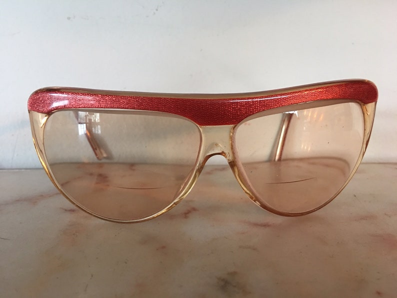 May include: A pair of vintage sunglasses with a red frame and clear lenses. The frame has a textured design.