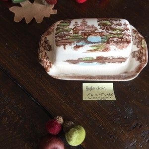 Puede incluir: Un plato de mantequilla de cer&aacute;mica blanca con una escena de paisaje marr&oacute;n y verde. El plato tiene un borde elevado y una peque&ntilde;a manija en el lateral. El plato mide 17,8 cm por 11,4 cm de ancho. El plato est&aacute; sobre una mesa de madera con una nota que dice "Butter Dish 7" x 4 1/2" wide bottom handle"