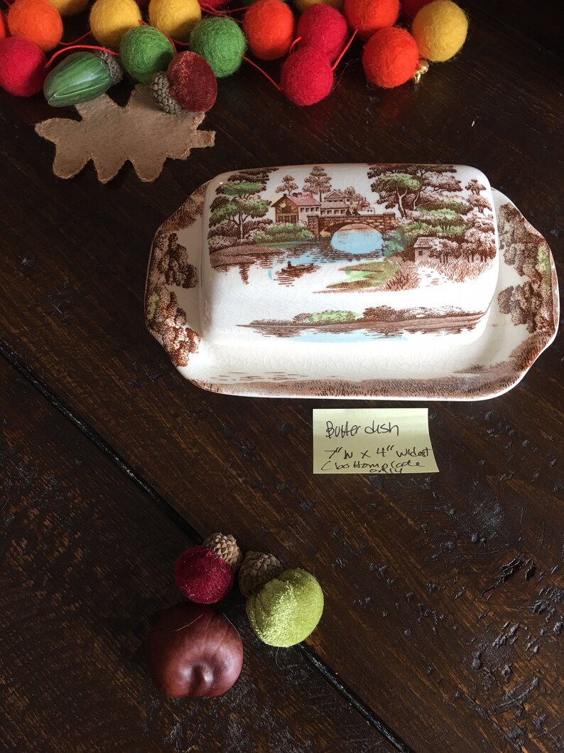 Puede incluir: Un plato de mantequilla de cer&aacute;mica blanca con una escena de paisaje marr&oacute;n y verde. El plato tiene un borde elevado y una peque&ntilde;a manija en el lateral. El plato mide 17,8 cm por 11,4 cm de ancho. El plato est&aacute; sobre una mesa de madera con una nota que dice "Butter Dish 7" x 4 1/2" wide bottom handle"