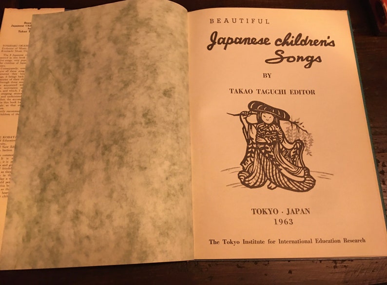 May include: The cover of a book titled "Beautiful Japanese Children's Songs" by Takao Taguchi Editor. The book cover features a black and white illustration of a child wearing a traditional Japanese outfit and holding a branch. The book was published in Tokyo, Japan in 1963 by The Tokyo Institute for International Education Research.