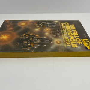 May include: A book cover with a gold spine and a black and white illustration of a futuristic city with stars in the background. The title of the book is "The Fall of Chronopolis" by Barrington J. Bayley. The book is part of the DAW Books series.