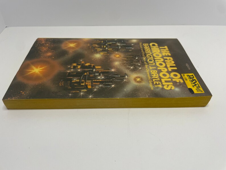 May include: A book cover with a gold spine and a black and white illustration of a futuristic city with stars in the background. The title of the book is "The Fall of Chronopolis" by Barrington J. Bayley. The book is part of the DAW Books series.