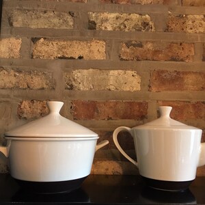 May include: Two white ceramic kitchenware items, a teapot and a covered casserole dish, both with black bases, are displayed on a shelf against a brick wall.