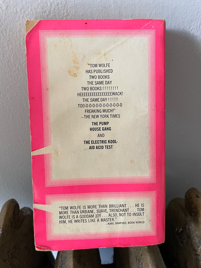 May include: A pink book cover with white text. The text reads "TOM WOLFE HAS PUBLISHED TWO BOOKS THE SAME DAY TWO BOOKS!!!!!!! HEEEEEEEEEEEEEEEWACK! THE SAME DAY!!!!! T00-0-0-0-0-0-0-0-0-0-0-0 FREAKING MUCH!" -THE NEW YORK TIMES THE PUMP HOUSE GANG AND THE ELECTRIC KOOL-AID ACID TEST "TOM WOLFE IS MORE THAN BRILLIANT... HE IS MORE THAN URBANE, SUAVE, TRENCHANT... TOM WOLFE IS A GODDAM JOY... ALSO, NOT TO INSULT HIM, HE WRITES LIKE A MASTER." -KARL SHAPIRO, BOOK WORLD