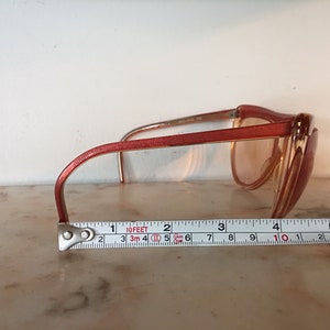 May include: A pair of pink plastic framed sunglasses with brown lenses. The sunglasses are sitting on a white surface with a measuring tape underneath.