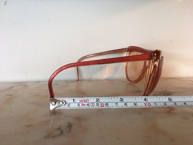 May include: A pair of pink plastic framed sunglasses with brown lenses. The sunglasses are sitting on a white surface with a measuring tape underneath.