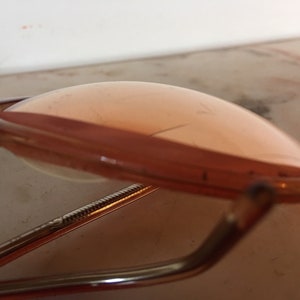 May include: A pair of vintage gold-tone glasses with pink tinted lenses. The glasses are folded in half and the lenses are facing up.
