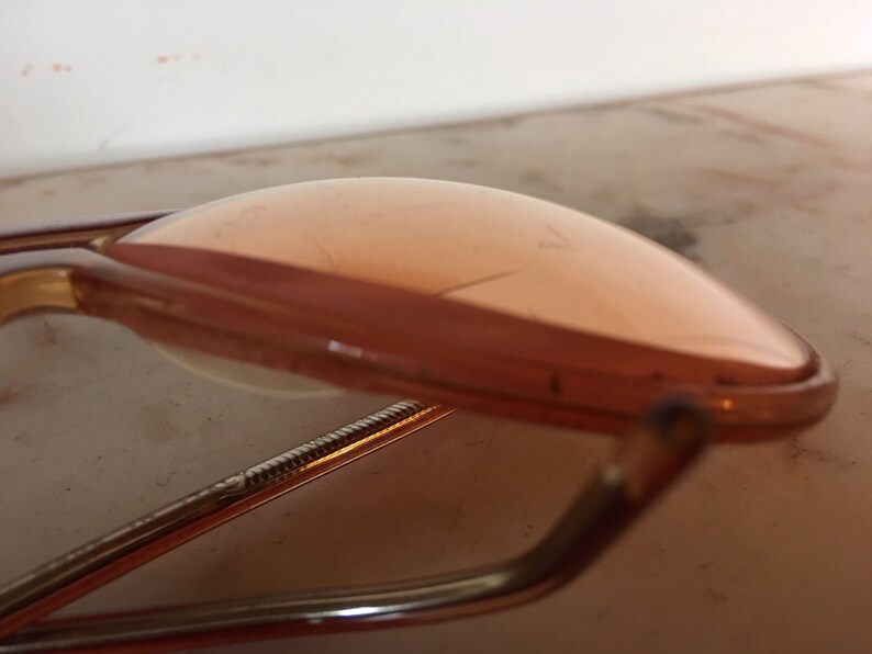 May include: A pair of vintage gold-tone glasses with pink tinted lenses. The glasses are folded in half and the lenses are facing up.