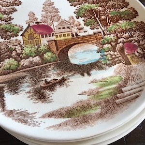 Puede incluir: Un juego de cuatro platos de cer&aacute;mica blanca con un dise&ntilde;o marr&oacute;n y verde de un puente sobre un r&iacute;o con un peque&ntilde;o bote en el agua. El dise&ntilde;o tambi&eacute;n incluye &aacute;rboles y casas.