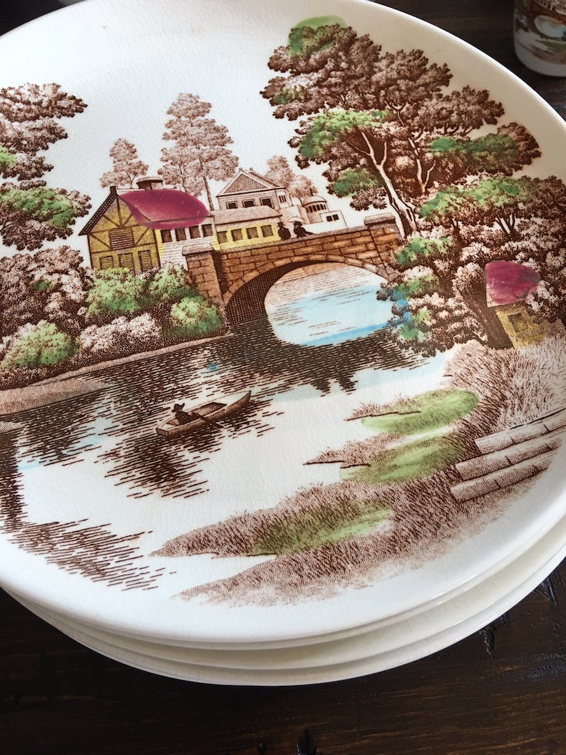 Puede incluir: Un juego de cuatro platos de cer&aacute;mica blanca con un dise&ntilde;o marr&oacute;n y verde de un puente sobre un r&iacute;o con un peque&ntilde;o bote en el agua. El dise&ntilde;o tambi&eacute;n incluye &aacute;rboles y casas.