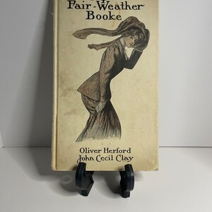May include: A vintage book titled "Cupid's Fair-Weather Booke" by Oliver Herford and John Cecil Clay. The cover features a black and white illustration of a woman in a long dress and hat, with her scarf blowing in the wind.