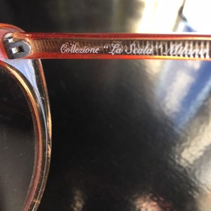 May include: Close-up of the arm of a pair of orange-tinted sunglasses with the text "Collezione "La Scala" Milano" etched into the frame.
