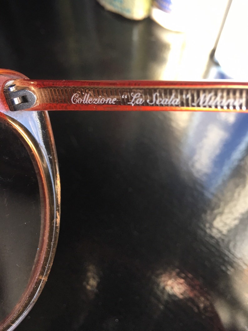 May include: Close-up of the arm of a pair of orange-tinted sunglasses with the text "Collezione "La Scala" Milano" etched into the frame.