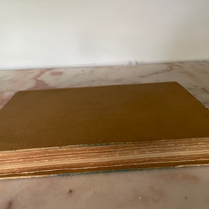 May include: A vintage book with a tan cover and aged, yellowed pages. The book's edges show wear, suggesting it's an antique. The cover is a solid color, and the pages are tightly bound.