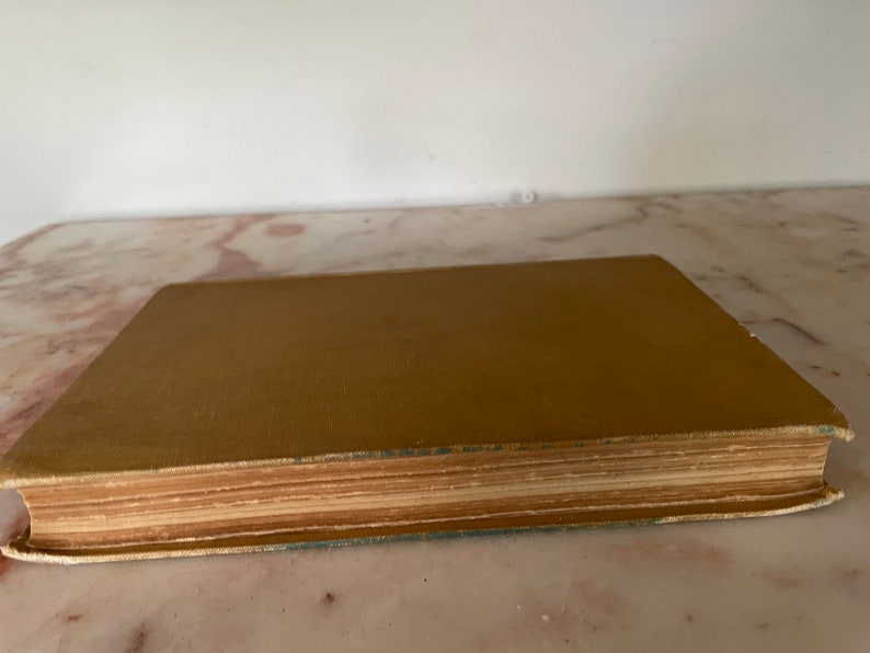May include: A vintage book with a tan cover and aged, yellowed pages. The book's edges show wear, suggesting it's an antique. The cover is a solid color, and the pages are tightly bound.