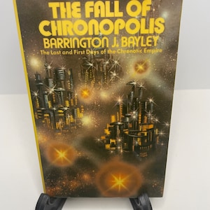 May include: A paperback book cover with a futuristic city scene in the background. The title is "The Fall of Chronopolis" by Barrington J. Bayley. The book is a DAW Books publication, number 105.