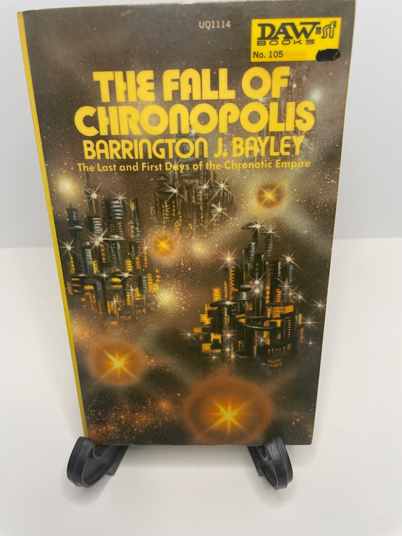 May include: A paperback book cover with a futuristic city scene in the background. The title is "The Fall of Chronopolis" by Barrington J. Bayley. The book is a DAW Books publication, number 105.