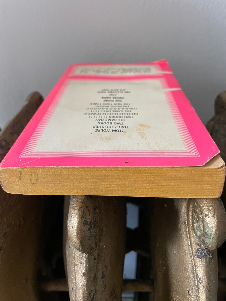 May include: Close-up of the spine of a pink paperback book with the text "TOM WOLFE HAS PUBLISHED TWO BOOKS THE SAME DAY" and "THE PUMP" printed on the spine.