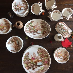 Puede incluir: Un juego de vajilla vintage de cer&aacute;mica marr&oacute;n y blanca con un dise&ntilde;o cottagecore que presenta un puente, &aacute;rboles y un r&iacute;o. El juego incluye platos, cuencos, tazas y una jarra.