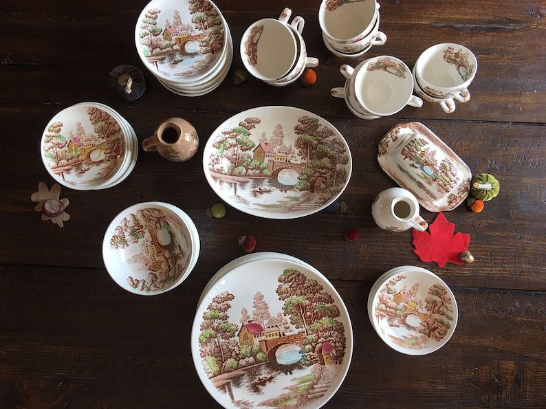 Puede incluir: Un juego de vajilla vintage de cer&aacute;mica marr&oacute;n y blanca con un dise&ntilde;o cottagecore que presenta un puente, &aacute;rboles y un r&iacute;o. El juego incluye platos, cuencos, tazas y una jarra.