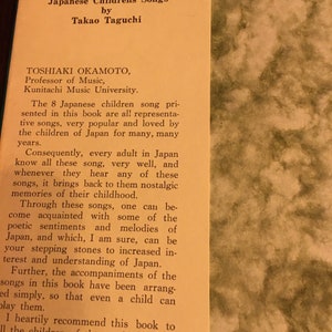 May include: A book cover with a yellow background and black text. The title is "Beautiful Japanese Children's Songs" by Takao Taguchi. The book is about Japanese children's songs and includes information about the author, Toshiaki Okamoto, Professor of Music at Kunitachi Music University.