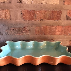 May include: A teal ceramic serving dish with a wavy edge and a light brown rim. The dish is sitting on a dark brown surface.