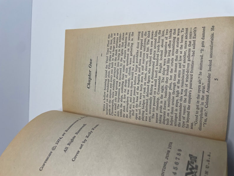 May include: A close-up of a book's open pages, showing the text "Chapter One" and the beginning of a story about a fleet of ships. The text describes the ships as being large and imposing, with a "porchlike door" opening on the flagship.