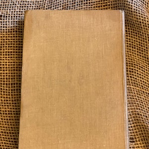 May include: A closed, rectangular book with a tan linen cover. The book's spine is visible on the right side, and the cover has a slightly textured appearance. The background is a woven, tan material.
