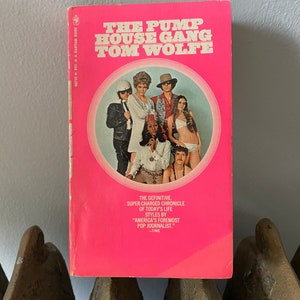 May include: A pink paperback book with a white circle on the cover. The circle contains a photo of six people. The book title is "The Pump House Gang" by Tom Wolfe. The text below the title reads "The definitive, super-charged chronicle of today's life styles by "America's foremost pop journalist." -TIME"