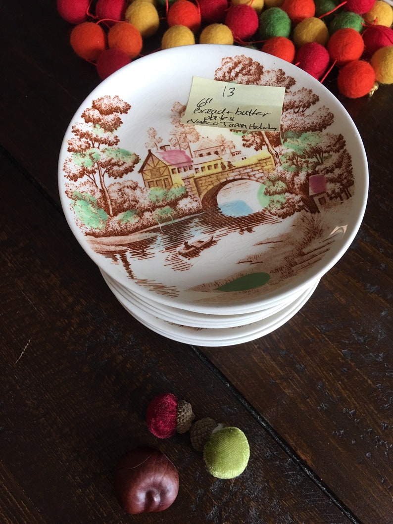 Puede incluir: Una pila de platos de pan y mantequilla de 15,24 cm de di&aacute;metro con un dise&ntilde;o floral marr&oacute;n y blanco que presenta un puente y una casa. Los platos est&aacute;n etiquetados con "6" Bread + butter plates, Nasco Town Holiday" en una nota adhesiva amarilla.
