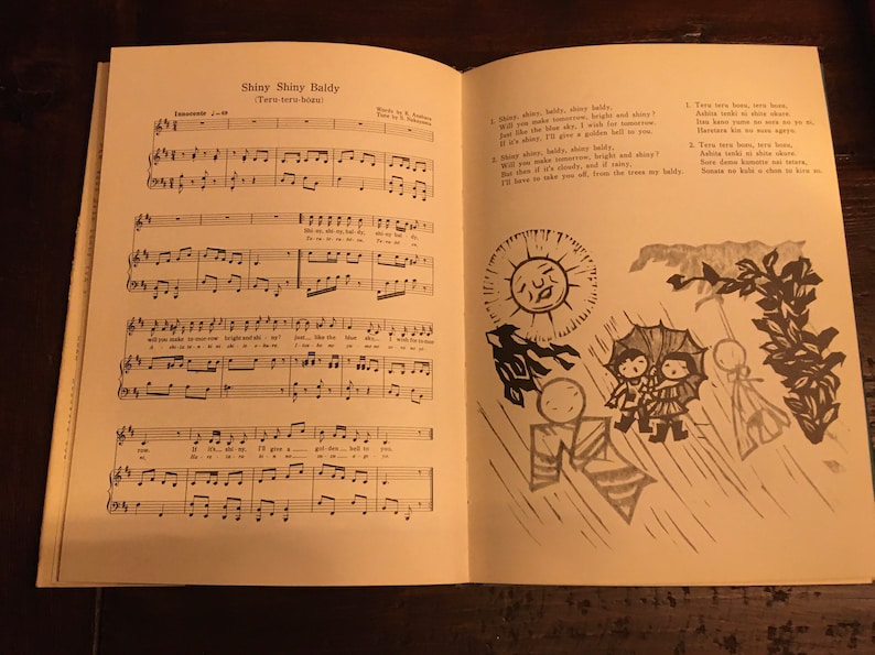 May include: A vintage sheet music book with a black and white illustration of a sun, rain, and people with umbrellas. The sheet music is for a song called "Shiny Shiny Baldy (Terr-terr-terr beat)".