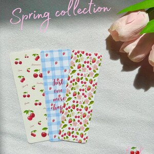 May include: Three bookmarks with a spring theme. The first bookmark has a white background with a repeating pattern of red cherries and green leaves. The second bookmark has a blue and white gingham pattern. The third bookmark has a white background with a repeating pattern of red strawberries, pink flowers, and green leaves. The text "Just one more chapter" is printed on the second bookmark.