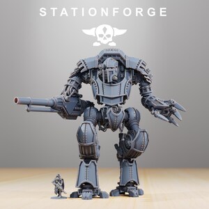 May include: A detailed grey model of a large war machine with a cannon and claw arm, next to a miniature soldier. The text "STATIONFORGE" and a skull logo are visible at the top.