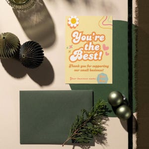 Puede incluir: Una tarjeta de agradecimiento amarilla con el texto "You're the Best!" en una fuente retro. La tarjeta tiene una margarita, corazones y remolinos de colores arco&iacute;ris. El texto "Thank you for supporting our small business!" est&aacute; debajo del texto principal. La tarjeta est&aacute; sobre una superficie beige con adornos verdes y un sobre verde.