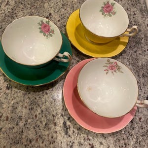 Op de afbeelding: Drie vintage theekopjes en schotels in groen, geel en roze. Elk theekopje is wit met een roze rozenontwerp. De schotels hebben een gouden rand. Een klassieke theeset.