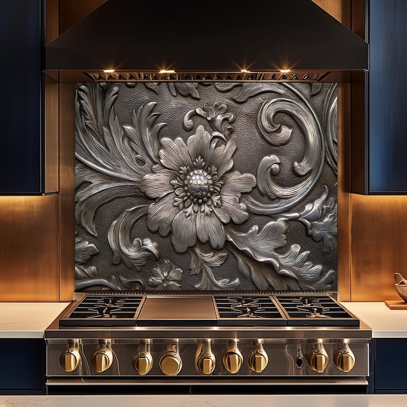 May include: A decorative kitchen backsplash with a large, detailed floral design in a metallic silver finish. The design features a central flower surrounded by leaves and swirling patterns. The backsplash is installed above a stainless steel gas range with gold knobs.