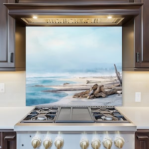 May include: A kitchen splashback displaying a tranquil beach vista. The image shows a sandy beach, driftwood, and a calm sea beneath a pale blue sky. The colour scheme is gentle, with blues, tans, and whites.