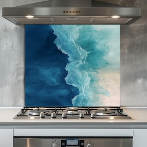 Könnte beinhalten: Eine Küchenrückwand aus Glas mit einem abstrakten Ozeanwellen-Design in Blau- und Weißtönen. Das Kunstwerk ist hinter einem Gasherd und unter einer Edelstahl-Dunstabzugshaube angebracht.