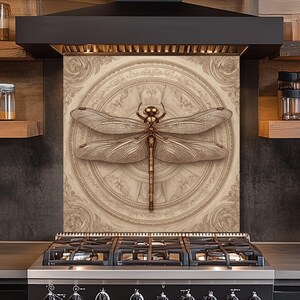 Peut inclure: Crédence de cuisine décorative avec un motif de libellule détaillé. La libellule est dans des tons marron et or, sur un fond beige avec un motif circulaire orné. La crédence est installée au-dessus d'une cuisinière à gaz noire.